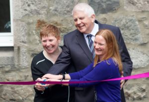 3 adults smiling while jointly holding a pair of scissors about to cut a ceremonial ribbon.